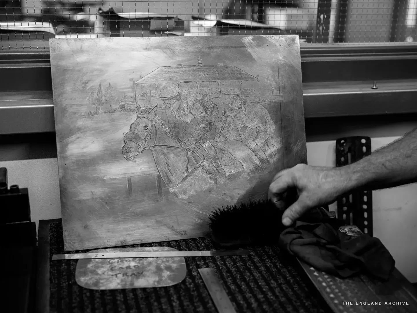 A hand brushes a large engraved steel plate with a horsehair brush, the plate etched with a horse-and-carriage coaching scene under a canopy.