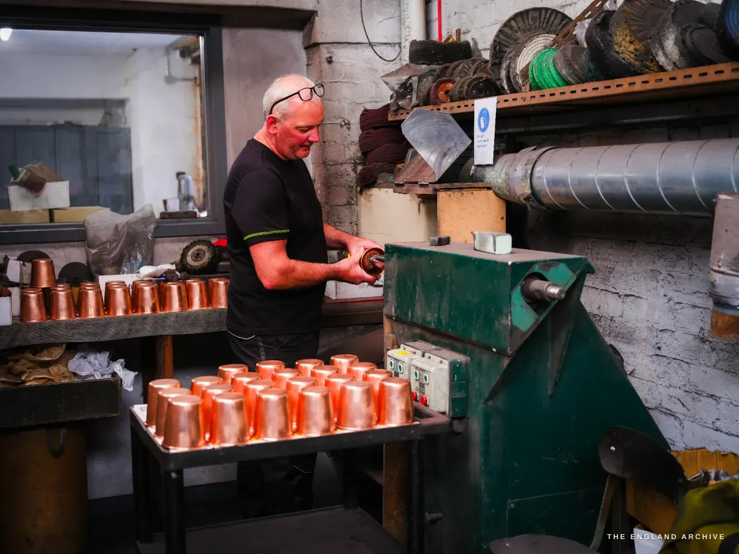 Phil at a green finishing machine, hands at the work, a tray of bright copper beaker blanks in the foreground, lagged ducting and safety notices on the wall.