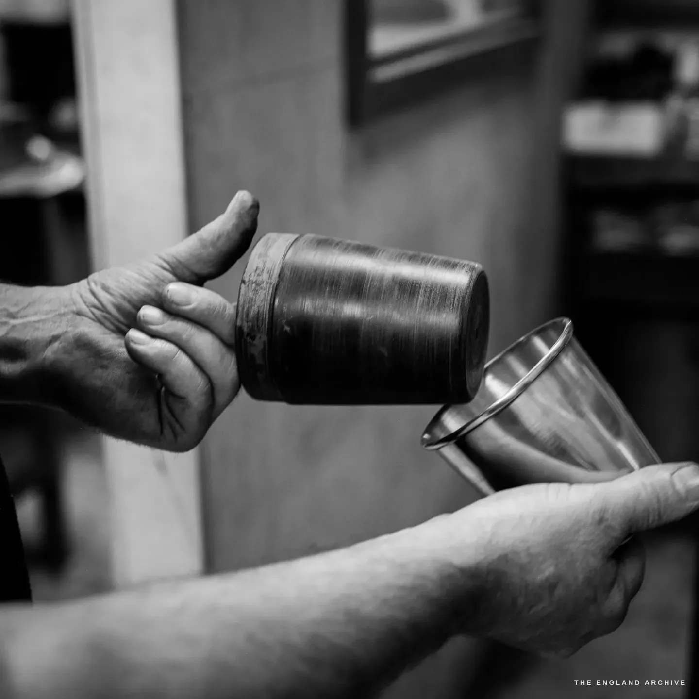 Two hands hold a dark cylindrical forming mould in the left and a bright finished metal beaker in the right, the beaker spun to shape over the mould.