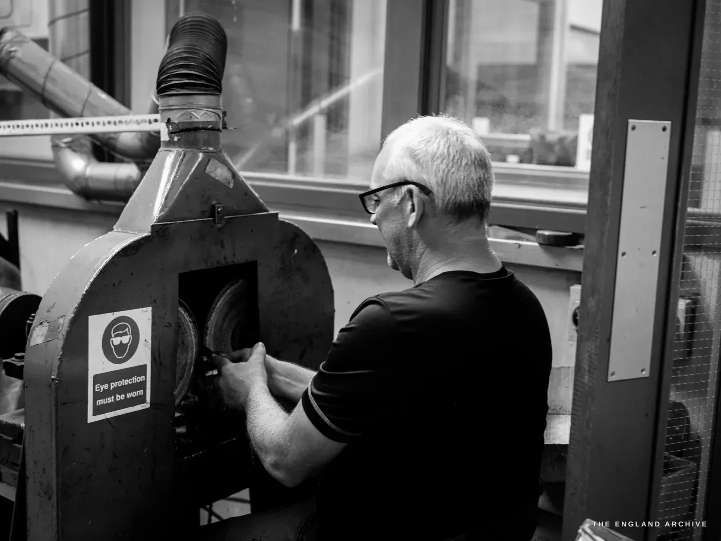Phil with his back to the camera working at the open mouth of a barrelling machine, a printed safety label on the housing reading Eye protection must be worn.
