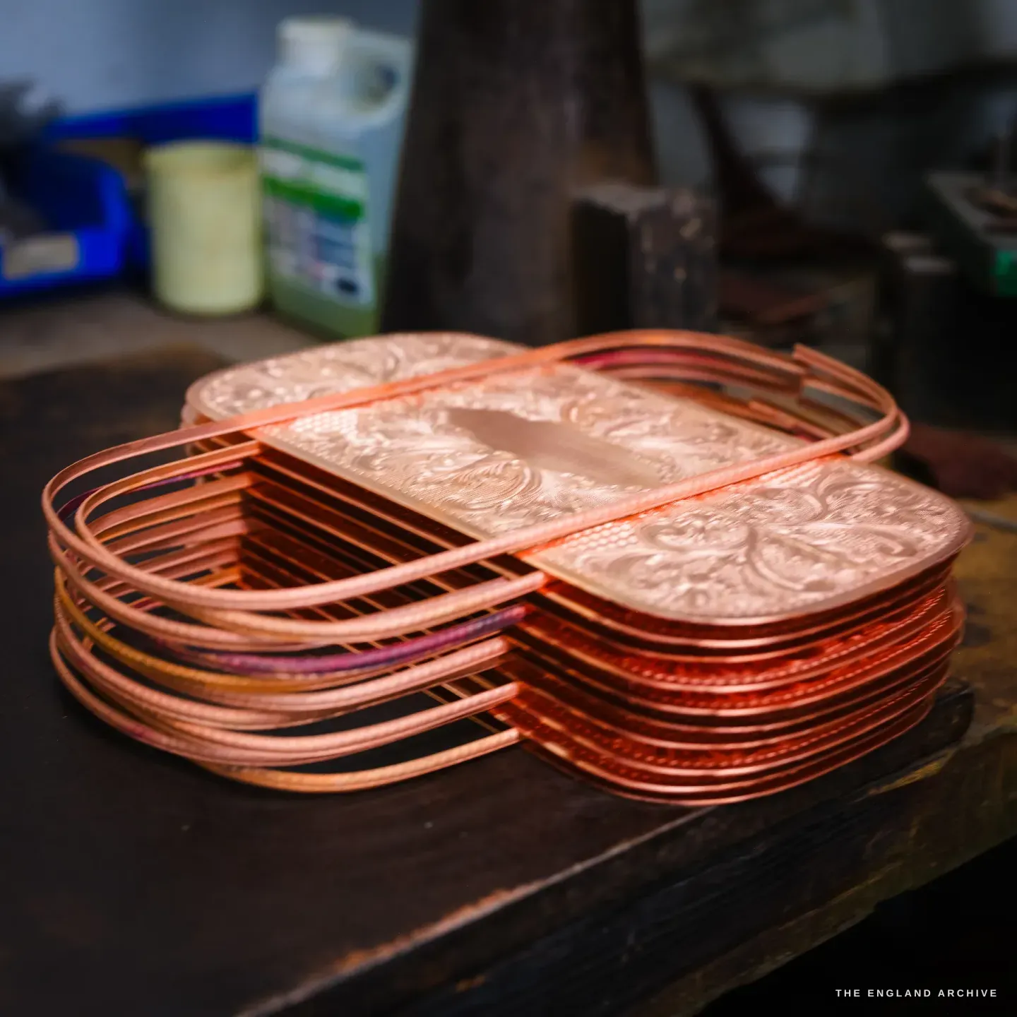 A stack of copper rounded-rectangular tray blanks on a dark bench, each embossed with dense floral scrollwork around a blank centre oval.