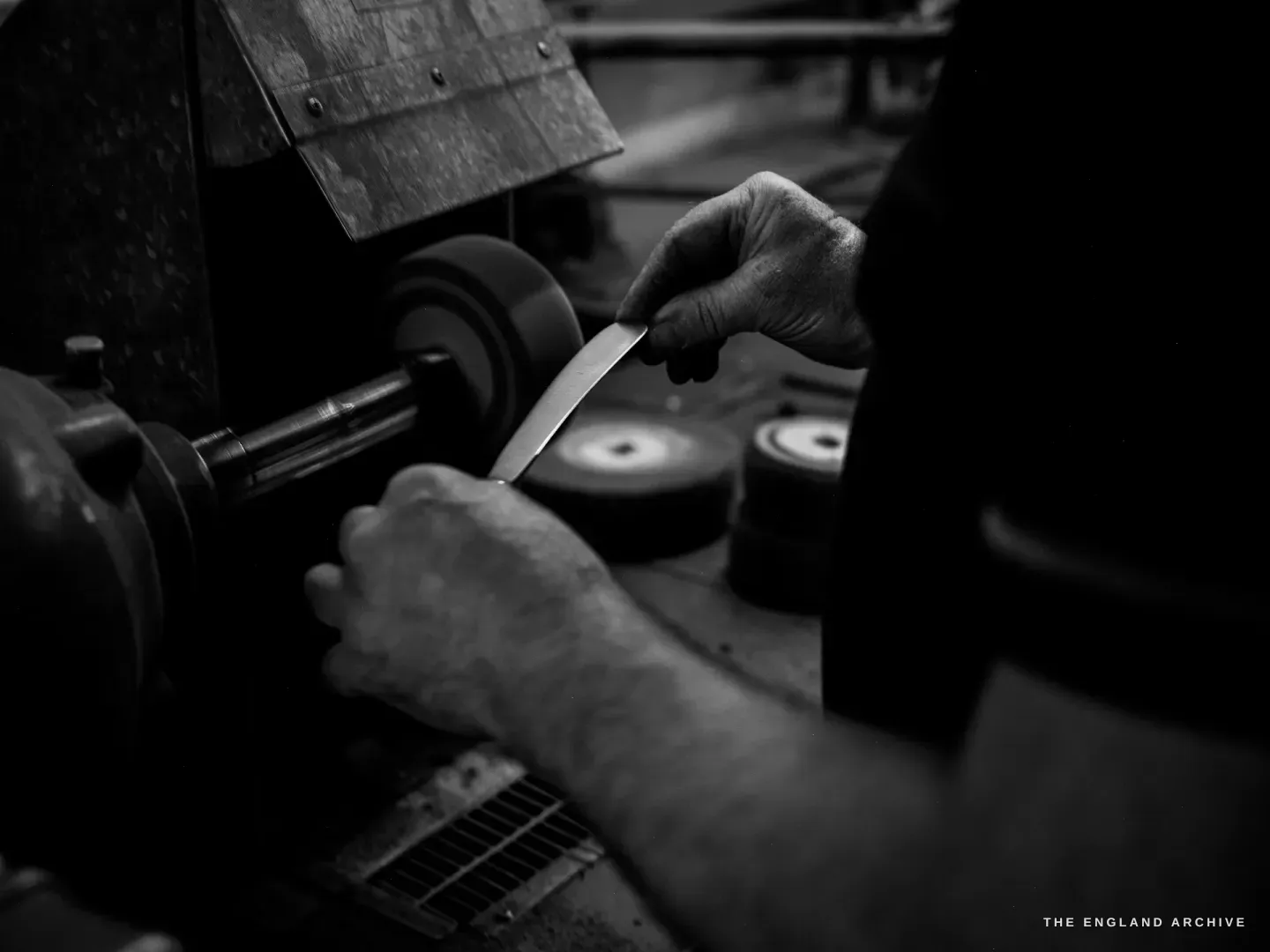 A low-key close-up of one hand pressing a thin curved blade against a small buffing wheel on a spindle, the other hand bracing below.