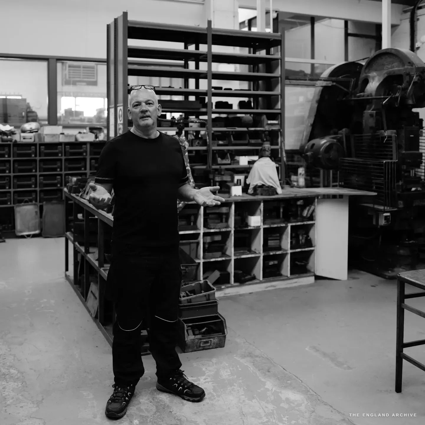 A full-length portrait of Phil standing in the centre of the workshop floor, one hand half-open in a gesture, a tall empty steel rack and a large press machine flanking him.