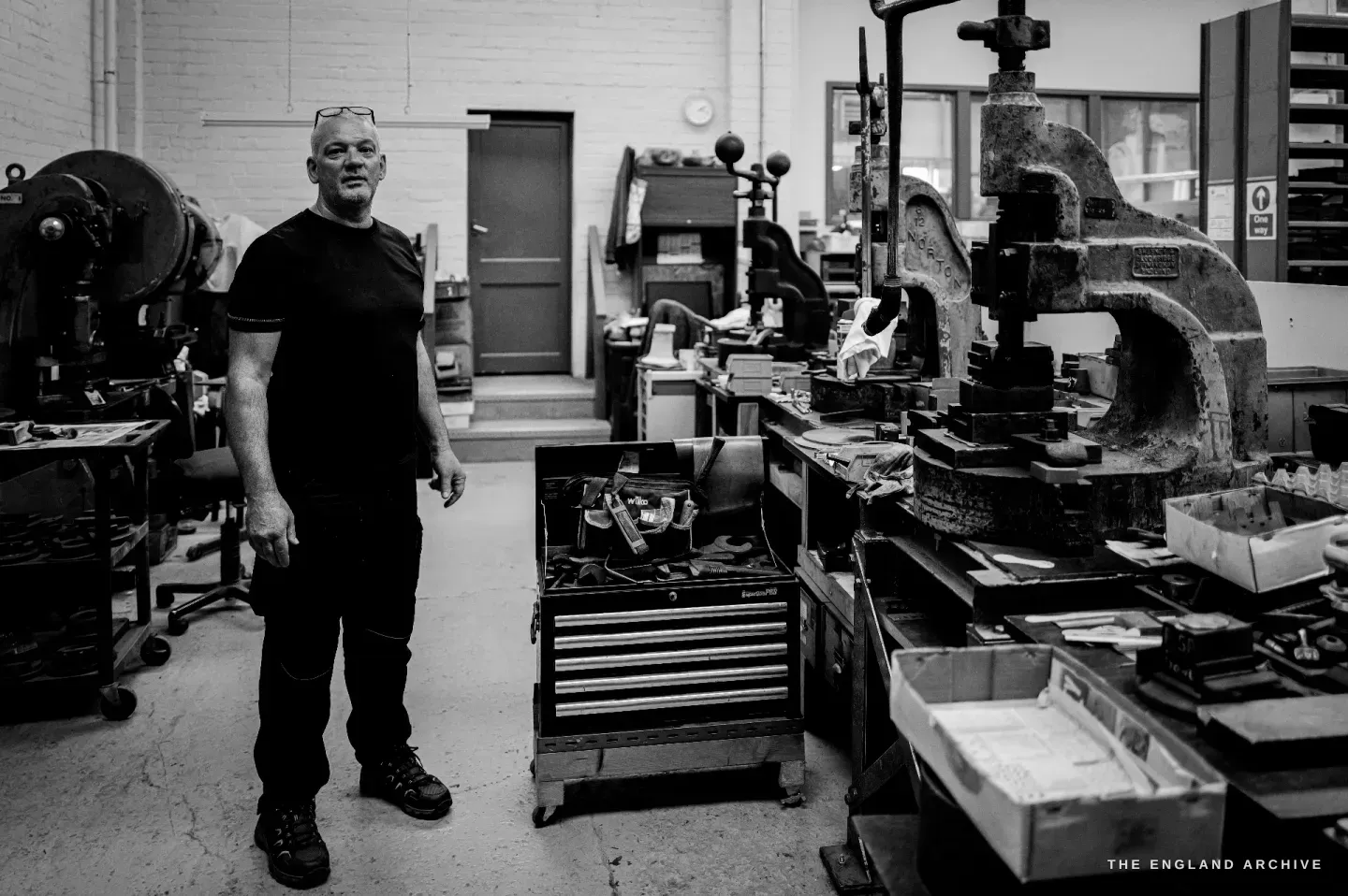 A full-length portrait of Phil standing relaxed beside a heavy cast fly-press, hand at his side, looking to camera, tool chests and benches around him.