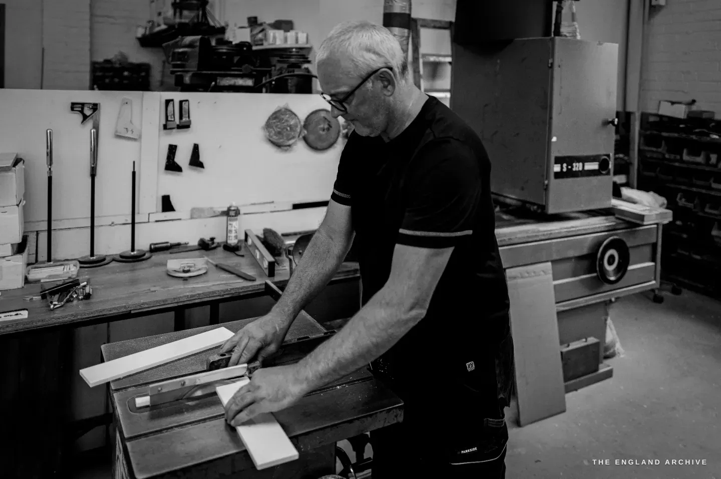 Phil feeding a long pale strip of stock across a bench machine, glasses on his nose, a tool board of templates on the wall behind him.
