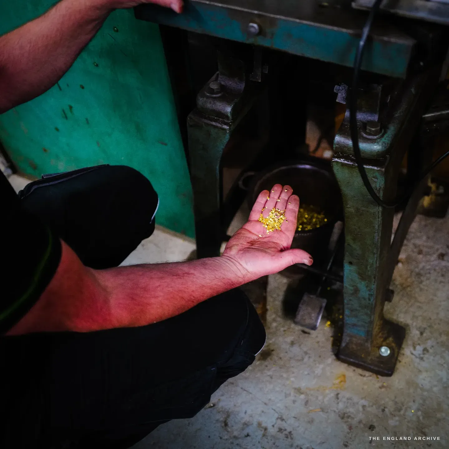 Phil crouched at a green clipping press, palm open to show a handful of small punched metal offcuts caught from beneath the press.