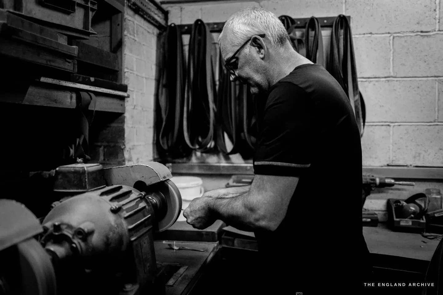 Phil in profile leaning into a bench polishing spindle, both hands at the wheel, rows of leather drive belts hung on the breeze-block wall behind him.