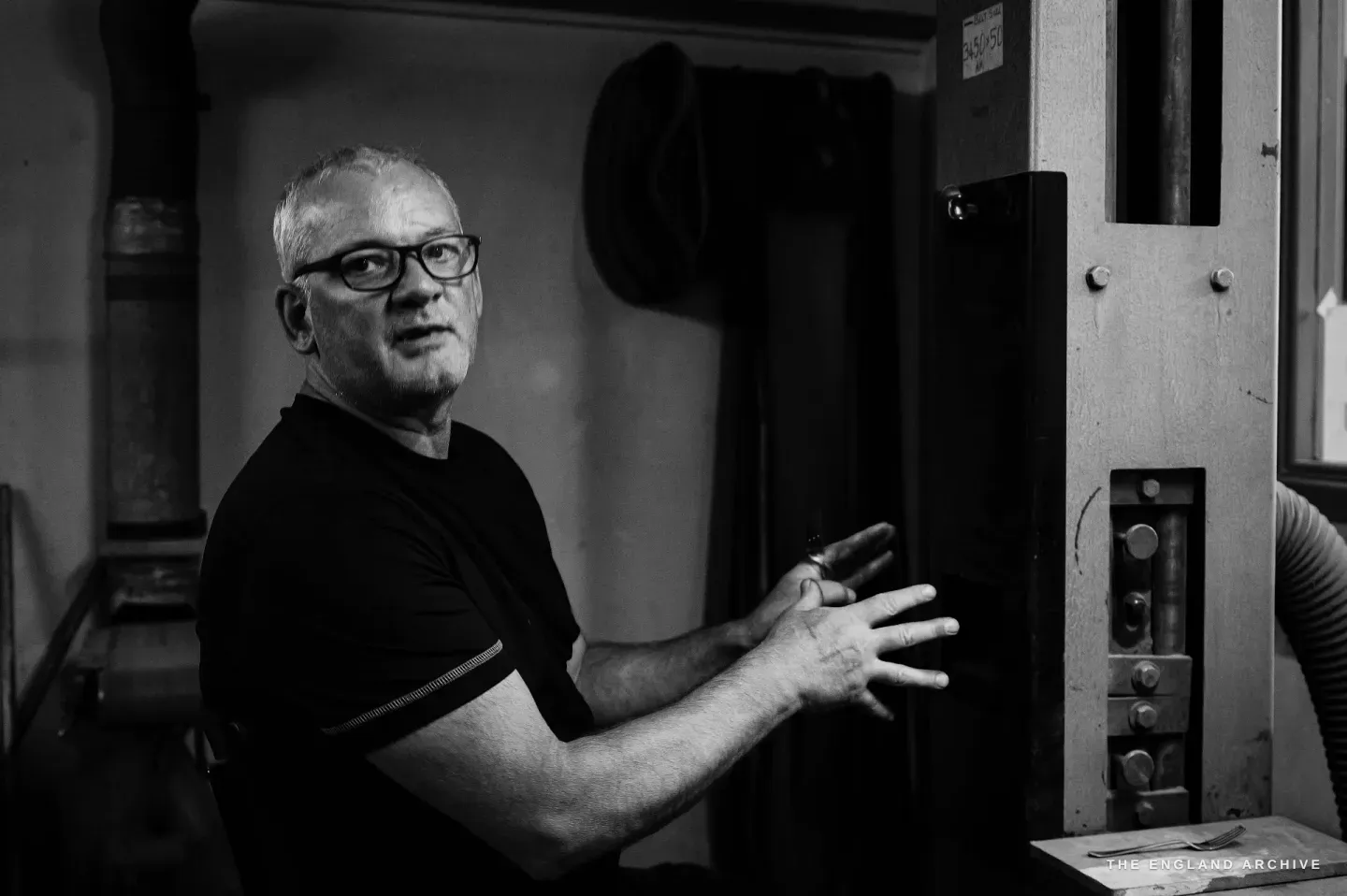 Phil half-turned to the camera, glasses on his nose, both hands raised mid-gesture as if explaining, a dark machine and hung belts behind him.