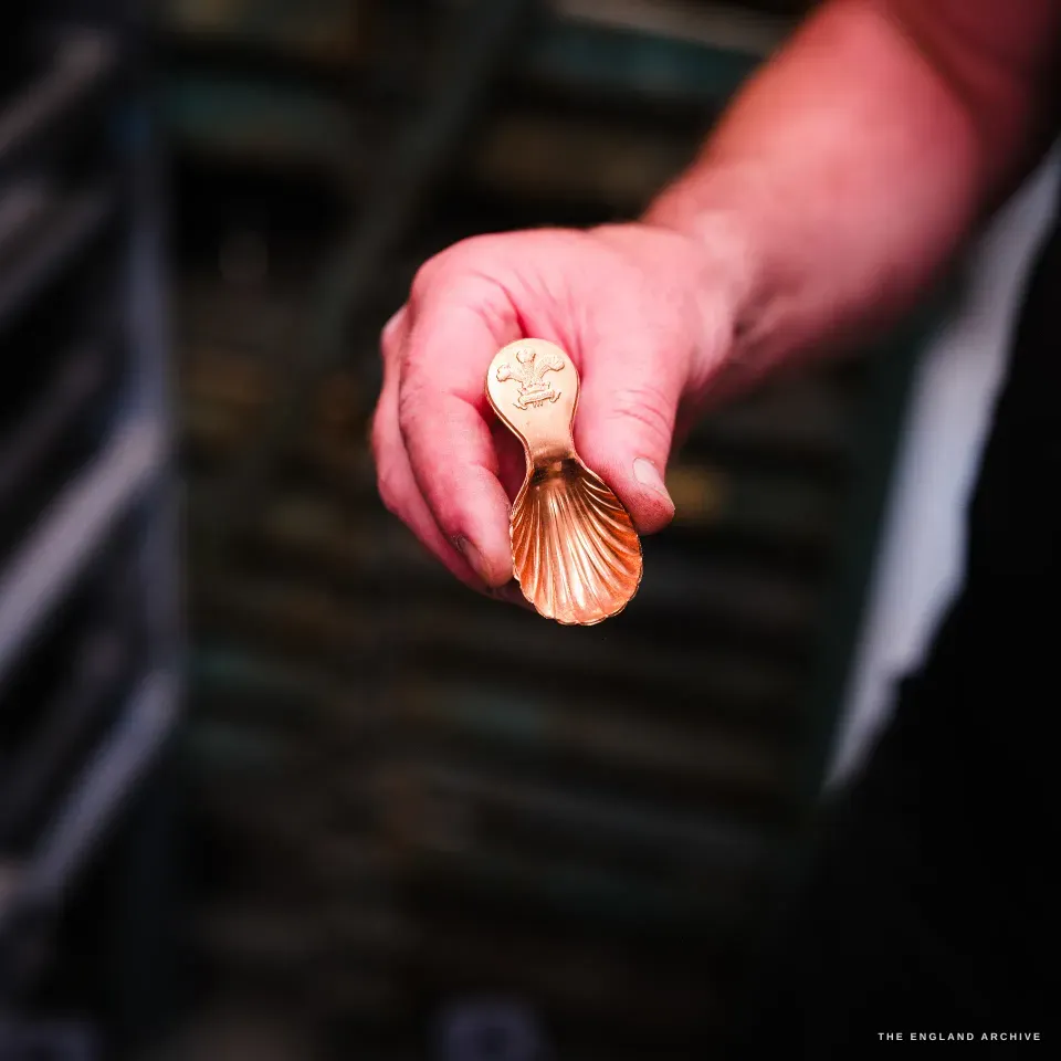 A hand holds out a small polished copper caddy spoon, its bowl fluted like a scallop shell, the stem stamped with the three-feathers Prince of Wales plumes crest.