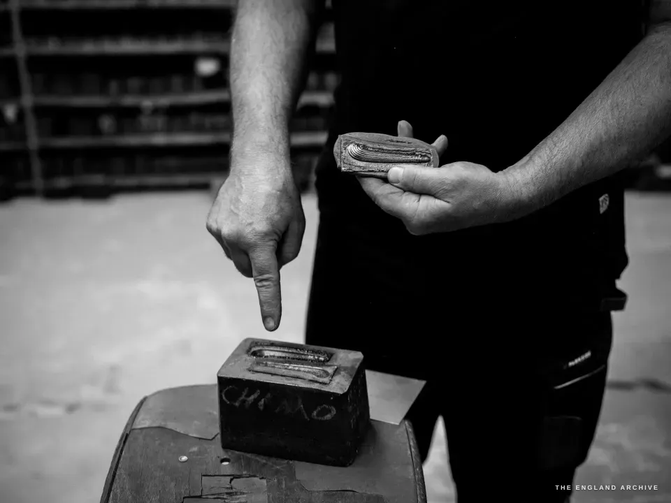 Phil, torso only, pointing down at one half of a two-part iron handle mould on a stool while holding the matching half in his other hand, the cavity shaped like a pistol grip.