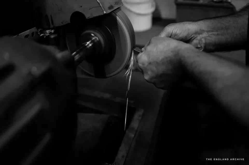 A tight low-key frame of Phil’s hands holding a thin metal strip to a fast-spinning buffing wheel, fine fibres of light streaming off the wheel.
