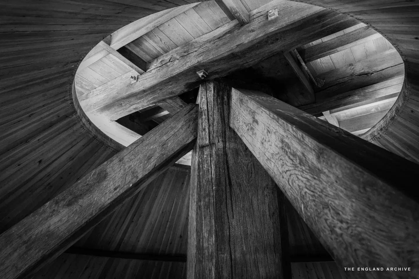 Looking straight up at the cross-frame of the mill: a circular timber rim with two heavy beams crossing through its centre, the boarded ceiling fanning out radially around them, every joint cut by hand.