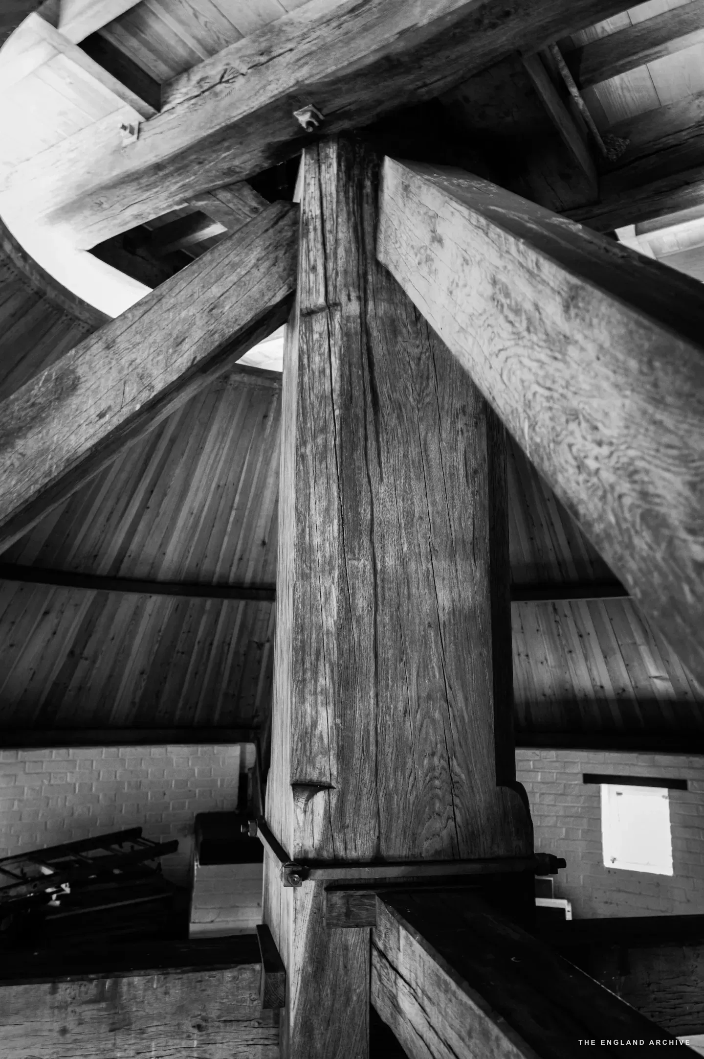 The central wooden mast of the post mill standing in the upper chamber: a single vertical column of enormous scale rising from the brick wall at the base to the timber-framed ceiling above, structural cross-beams radiating outward from it.