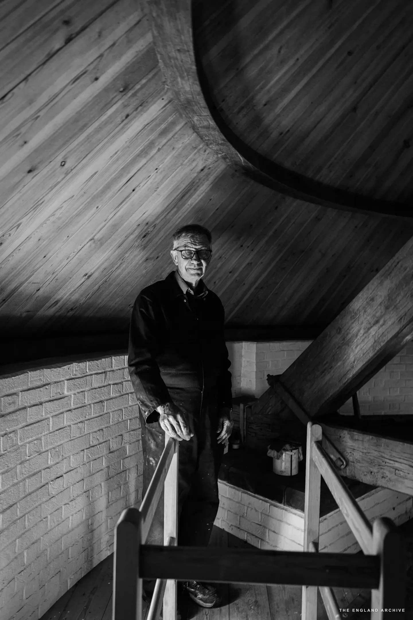 Richard standing at the head of the wooden staircase inside the upper chamber of the mill, the curved tongue-and-groove ceiling sweeping up and away above him, brick walls below.