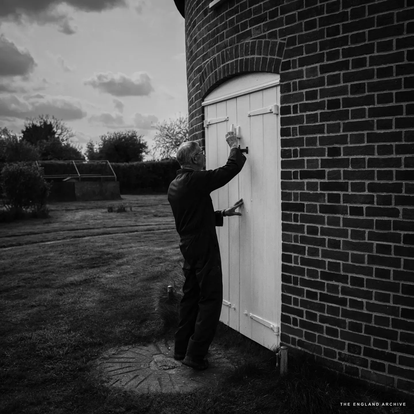 Richard Seago in his work overalls at the white wooden door set into the brick base of the post mill, reaching up to unlatch it.