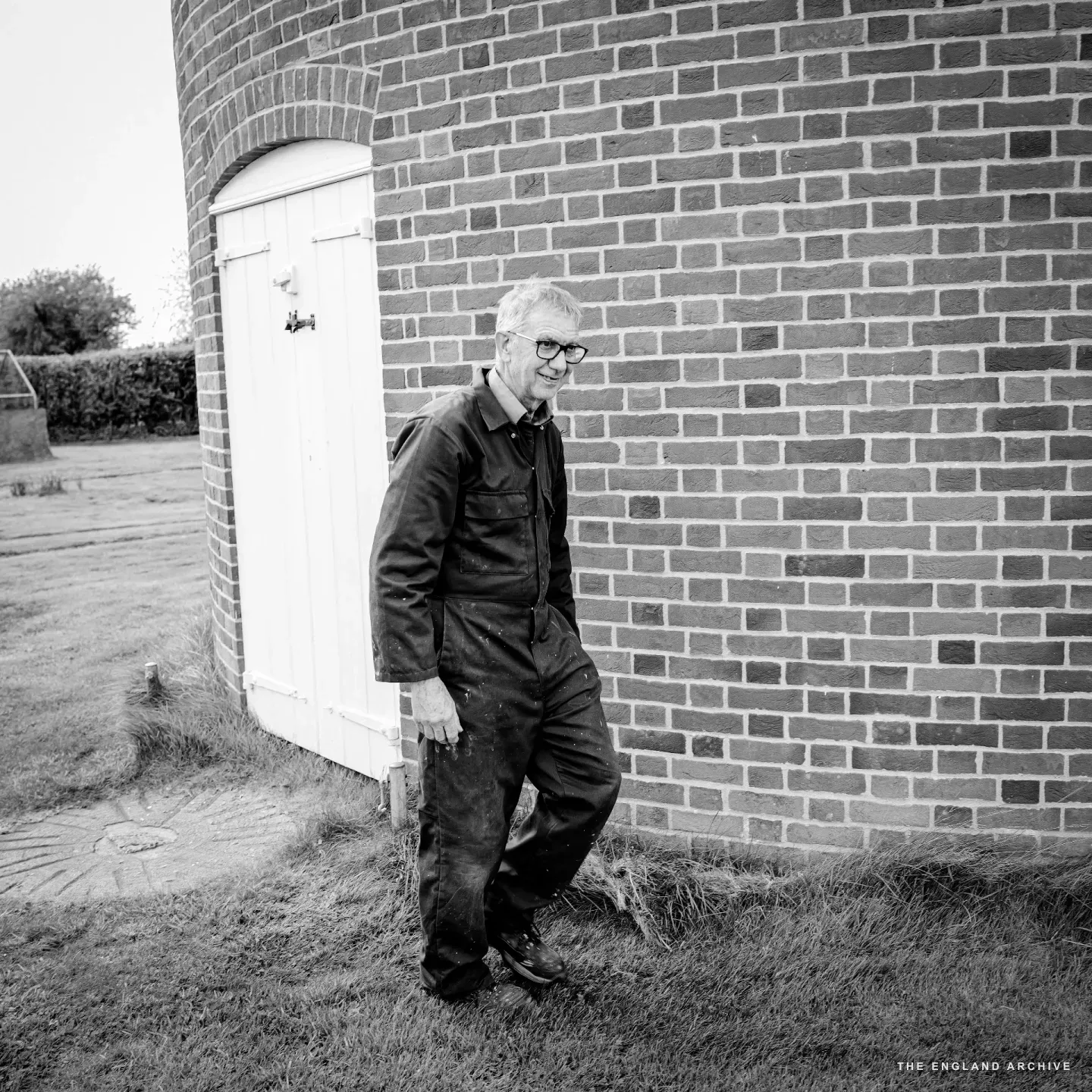 Richard Seago leaning easily against the brick wall of the roundhouse, hands in pockets, the white door behind him, looking towards the camera.