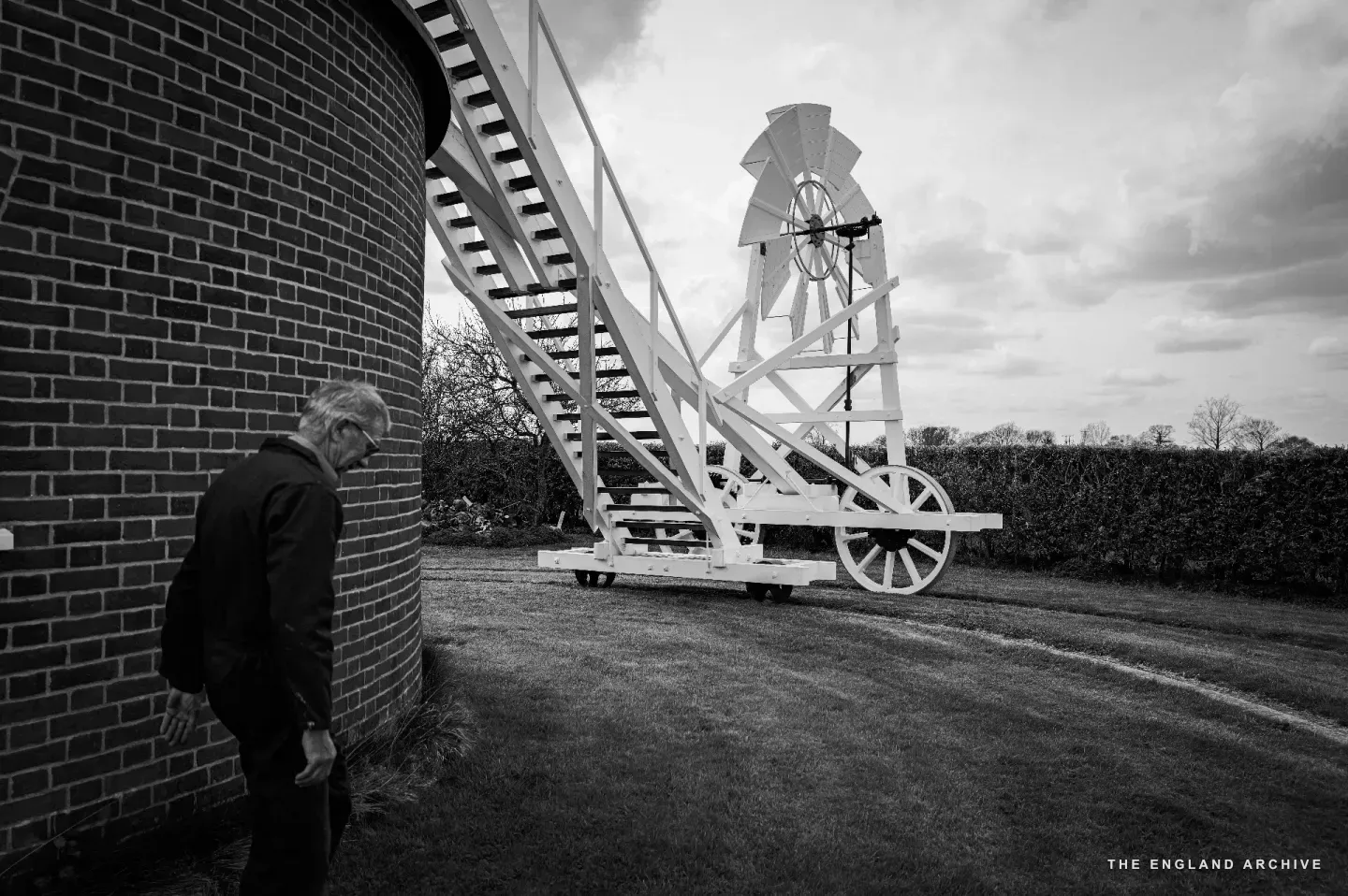 Richard standing in the foreground at the brick base of the mill, looking up at the white-painted fantail trolley: a stair-and-ladder structure on wheels, rising at an angle to a small wind-bladed fantail that turns the cap of the mill into the wind.