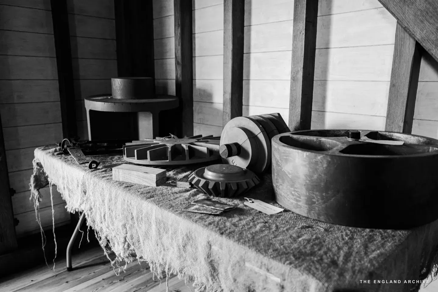 A bench in the woodworking shop with several wooden gear blanks, pulleys, and pattern parts laid out across a length of sacking, the whitewashed timber walls of the shop behind.