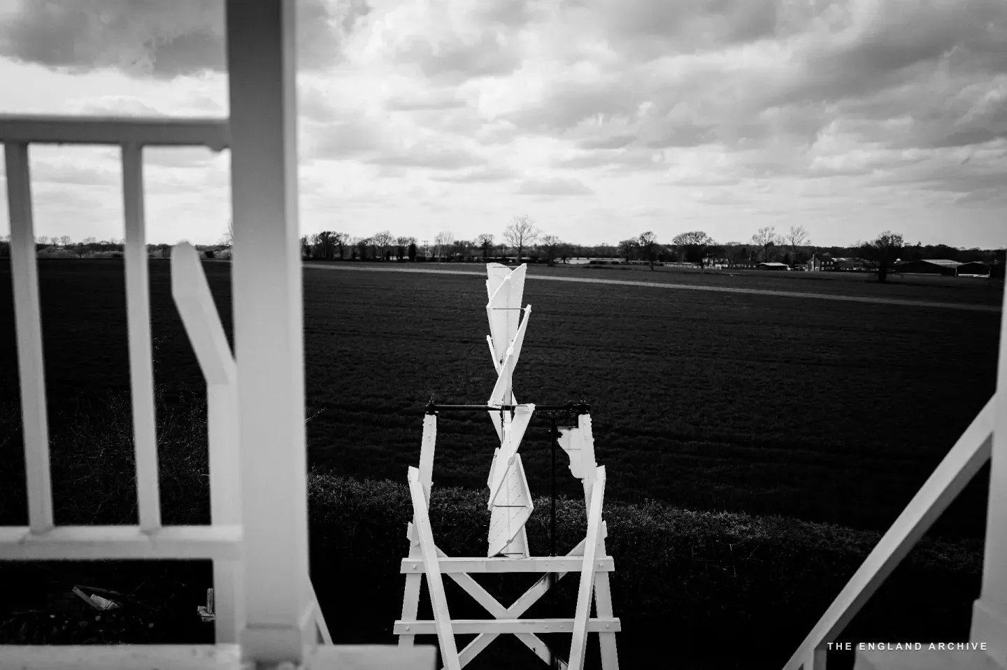 Looking out from the upper platform of the post mill through the white-painted railings: a small white iron fantail blade rising in the centre of the frame, beyond it the dark flat Norfolk fields running to a low wooded horizon.
