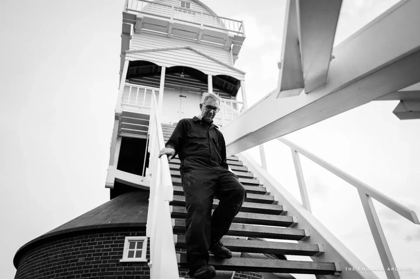 Richard descending the wooden staircase from the cap of the post mill, the white-clapboard upper body and the brick roundhouse curving away above and behind him, late afternoon light flat across the timber.