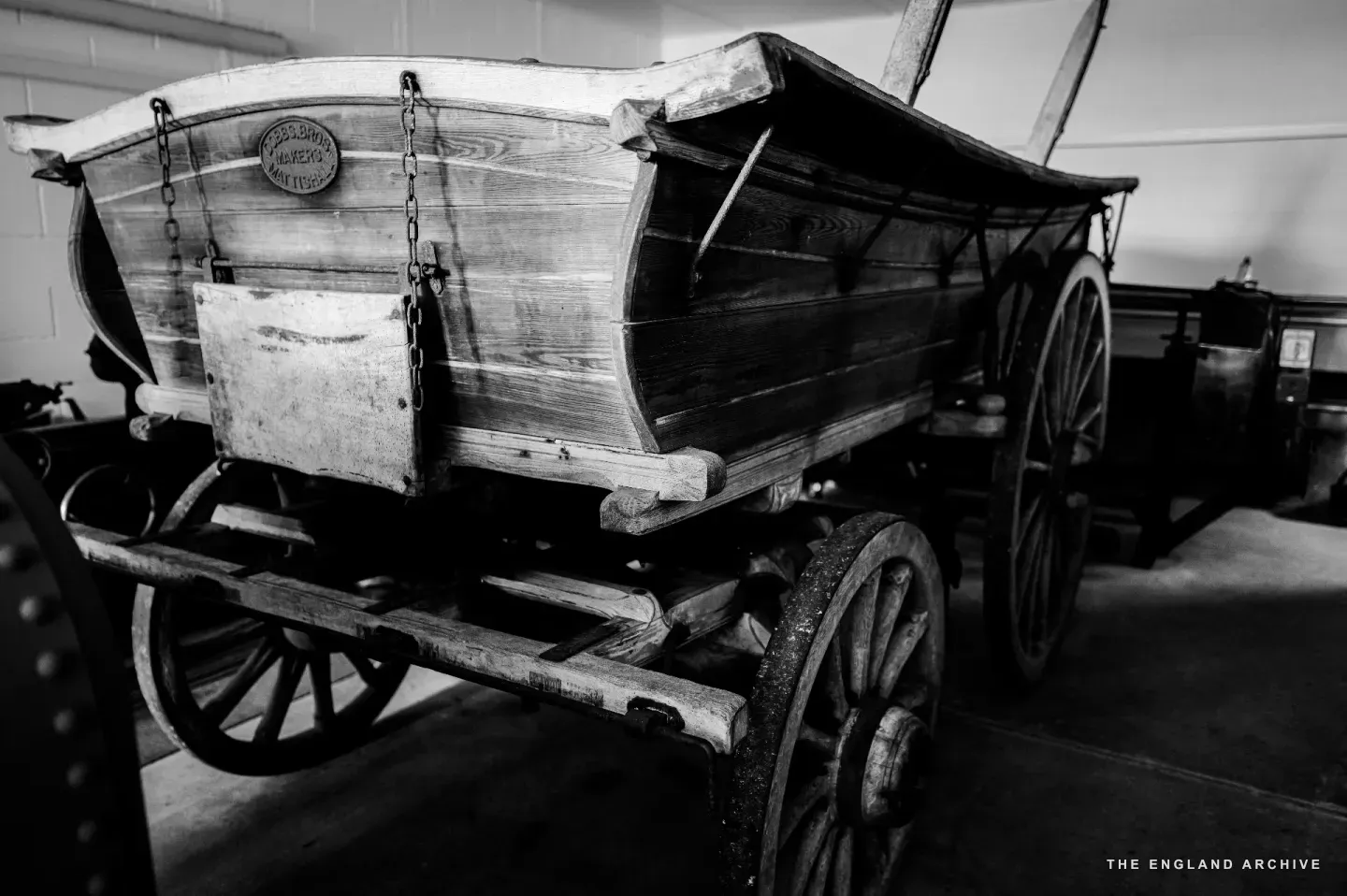 A restored horse-drawn farm wagon in the barn, photographed three-quarters from behind: tight-jointed timber sides, iron-rimmed wooden wheels, a small maker’s plate fixed to the headboard.