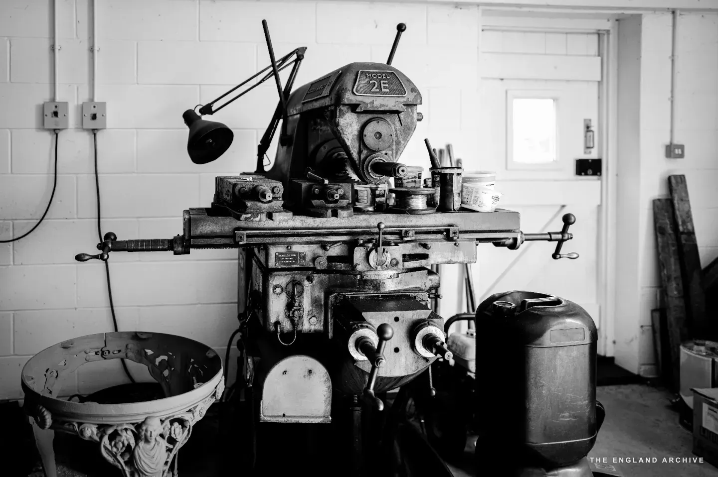 A vintage milling machine in the metalwork shop, the maker’s name MOSSEN above the spindle and the model number 2E plate on the casing, with hand-wheels, levers, and a small task lamp pulled in close.