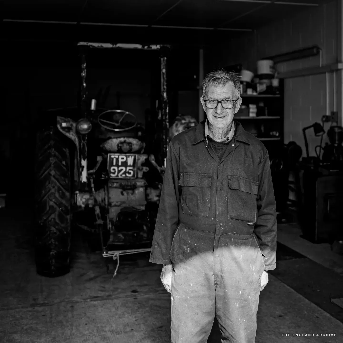 Richard standing inside the barn doorway in his work jacket and white trousers, hands at his sides, with a Fordson tractor registered TPW 925 parked in the dim space behind him to the left.