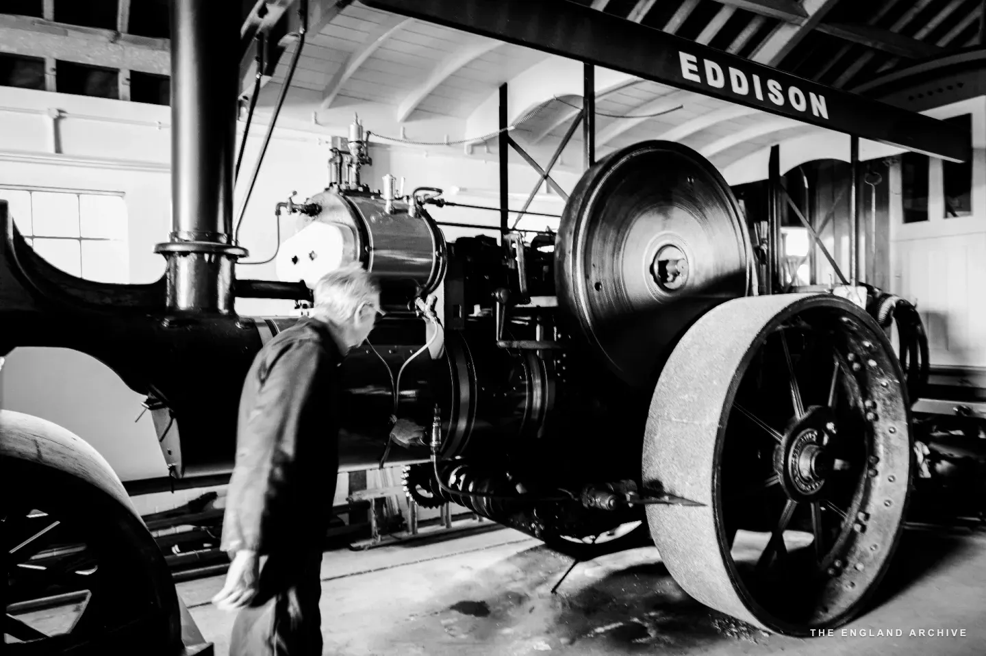 Richard standing alongside an Eddison steam roller in a long shed, the painted EDDISON sign visible on the rear wall above, the roller’s boiler, chimney, and tall driving wheel rising above him.