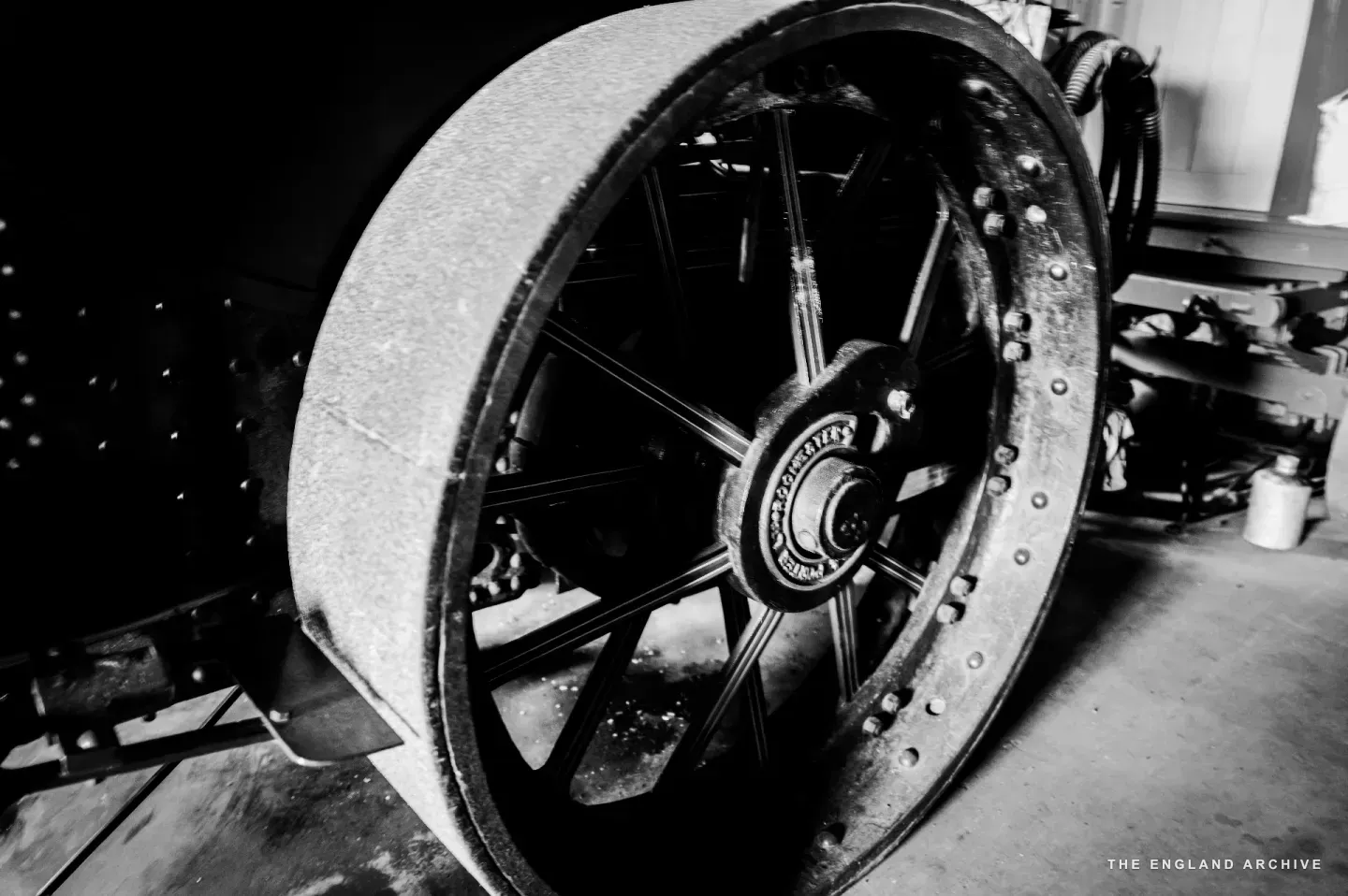 A close detail of the rear flywheel of the Eddison steam roller, photographed from low and side-on: the cast-iron rim, the spoked centre carrying the maker’s mark, the heavy bolted axle running through it.