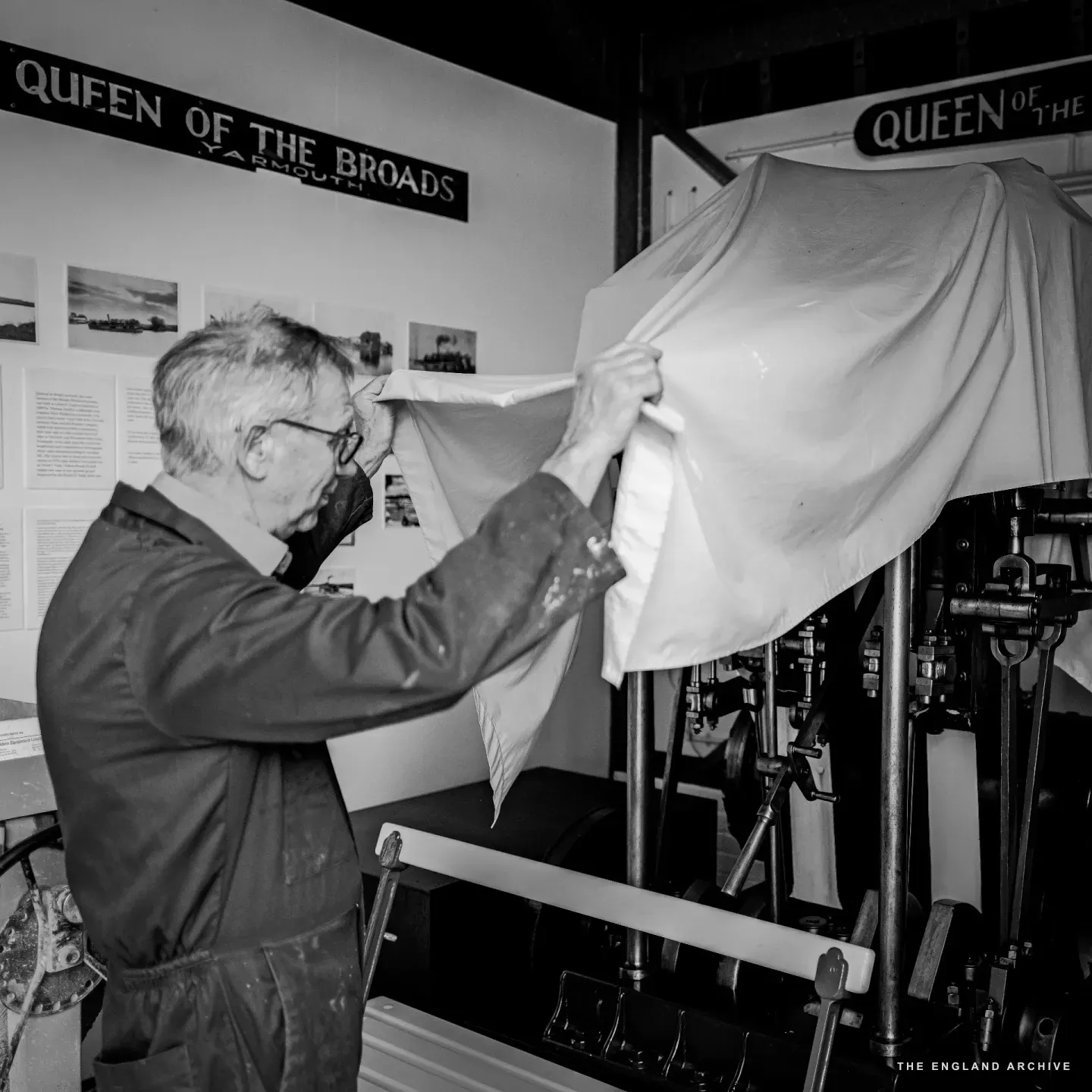 Richard pulling a white sheet up off the Queen of the Broads’ Victorian steam engine, the painted QUEEN OF THE BROADS / YARMOUTH signs from the original riverboat fixed to the wall above the engine.