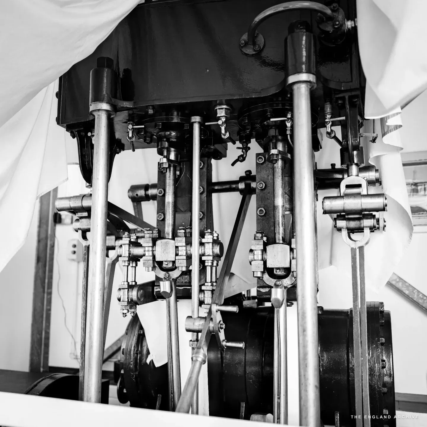 The exposed mechanism of the Queen of the Broads’ steam engine: three vertical polished cylinders, brass valves and rods running between them, the engine head and flywheel below, the white sheet still draped to either side.