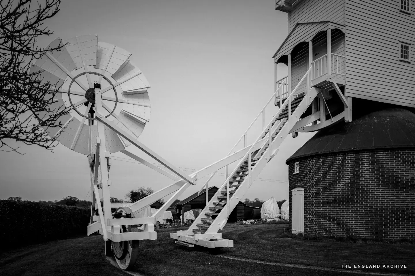 Side view of the post mill in late light, the long white fantail beam running diagonally from the wheeled trolley up to the back of the cap, the small bladed fantail wheel at its far end.