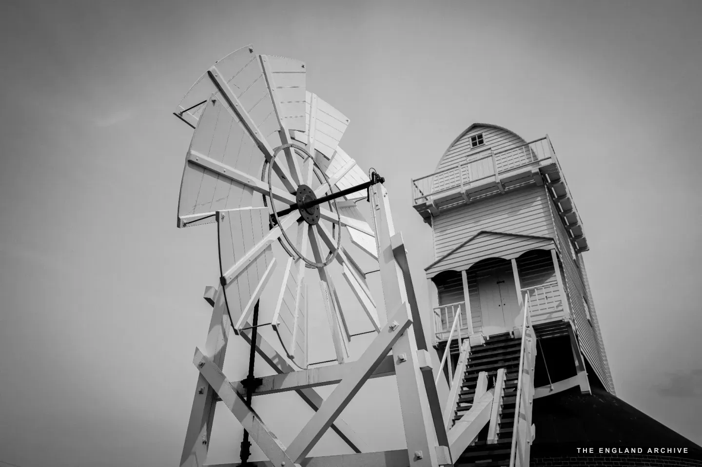 A close upward view of the fantail blades and the cap of the mill together against a pale cloudy sky, the wind-bladed wheel circular at left, the white-clapboard cap rising at right.