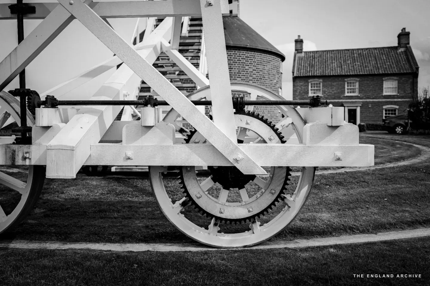 A low side-on view of the geared wheels at the base of the fantail trolley - cast iron toothed wheels and white timber framing - with the brick farmhouse Richard built standing in the background to the right.