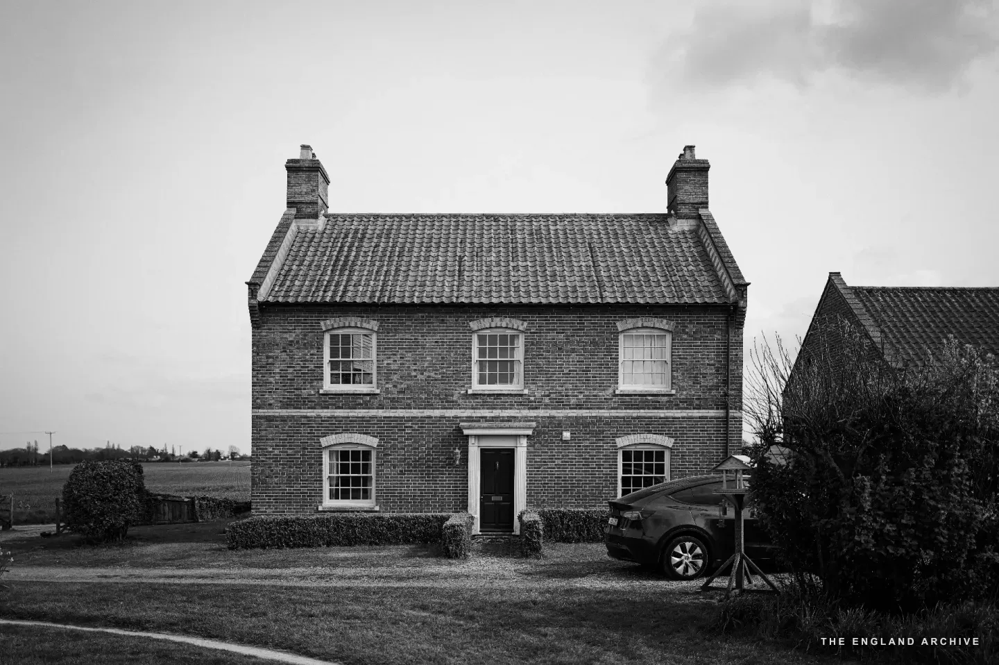 A red-brick two-storey farmhouse photographed front-on across a gravelled yard: pantile roof, two chimneys, sash windows, a small porch over the front door, an outbuilding to the right.