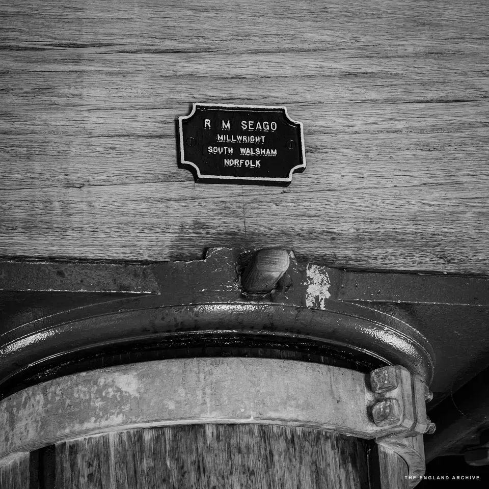 A small black-and-white maker’s plate fixed to the inside of the mill, reading R M SEAGO / MILLWRIGHT / SOUTH WALSHAM / NORFOLK, mounted on a length of pale weathered timber above the curved iron rim and round wooden base of the grindstone assembly.