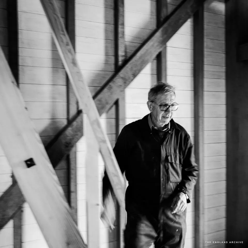 Richard descending a wooden staircase inside the woodworking shop, side-on, the diagonal beams of the stair-frame in the foreground and the white-painted tongue-and-groove walls behind.