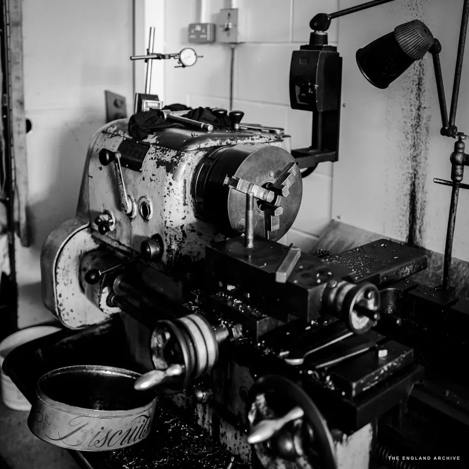 A bench lathe in the metalwork shop seen from a low three-quarter angle, the chuck and tool-post in sharp focus, swarf scattered along the bed, and a small Biscuits-marked tin sitting in the foreground catching the off-cuts.