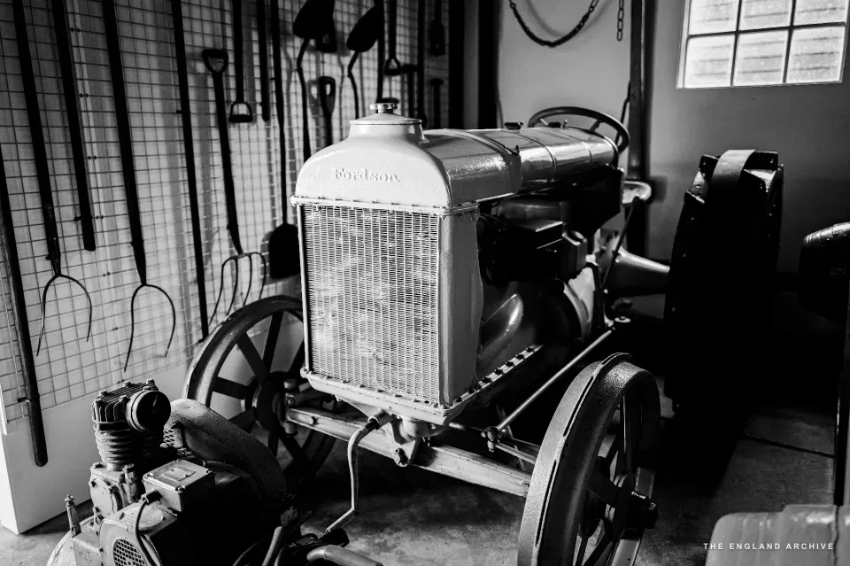 A 1920s Fordson tractor inside the barn, side-on, the radiator and bonnet bright in the available light, hand tools - billhooks, pitchforks, hayrakes - hanging on the wall behind.