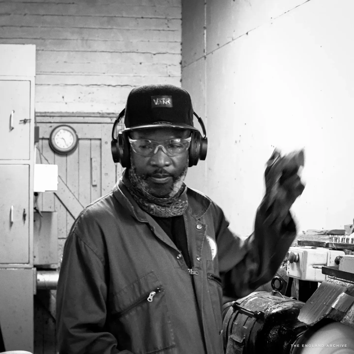 Sabino Henda in a cap, ear defenders and safety glasses with a scarf at his neck, a blackened glove raised, standing at his polishing station, a clock on the wall behind.