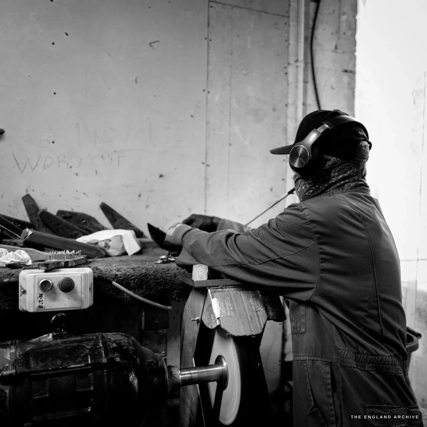 Sabino seen from behind at the polishing bench, leaning into the work, the buffing wheel and motor below, part-finished blades racked along the bench to his left.