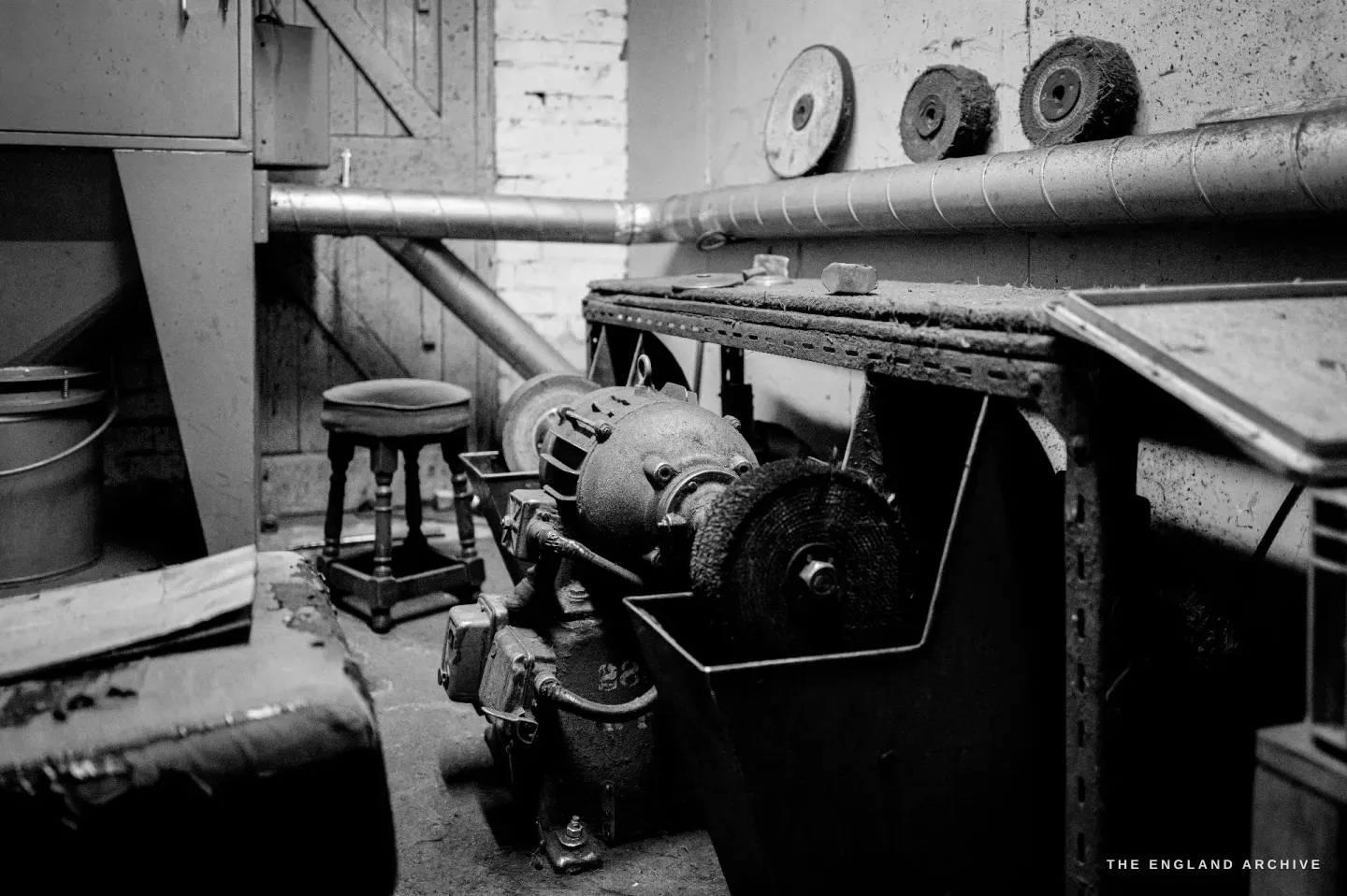 The polishing machine standing empty: buffing and abrasive wheels on the spindle, the motor and drive below, spare polishing mops hanging on the wall above, a stool to one side.