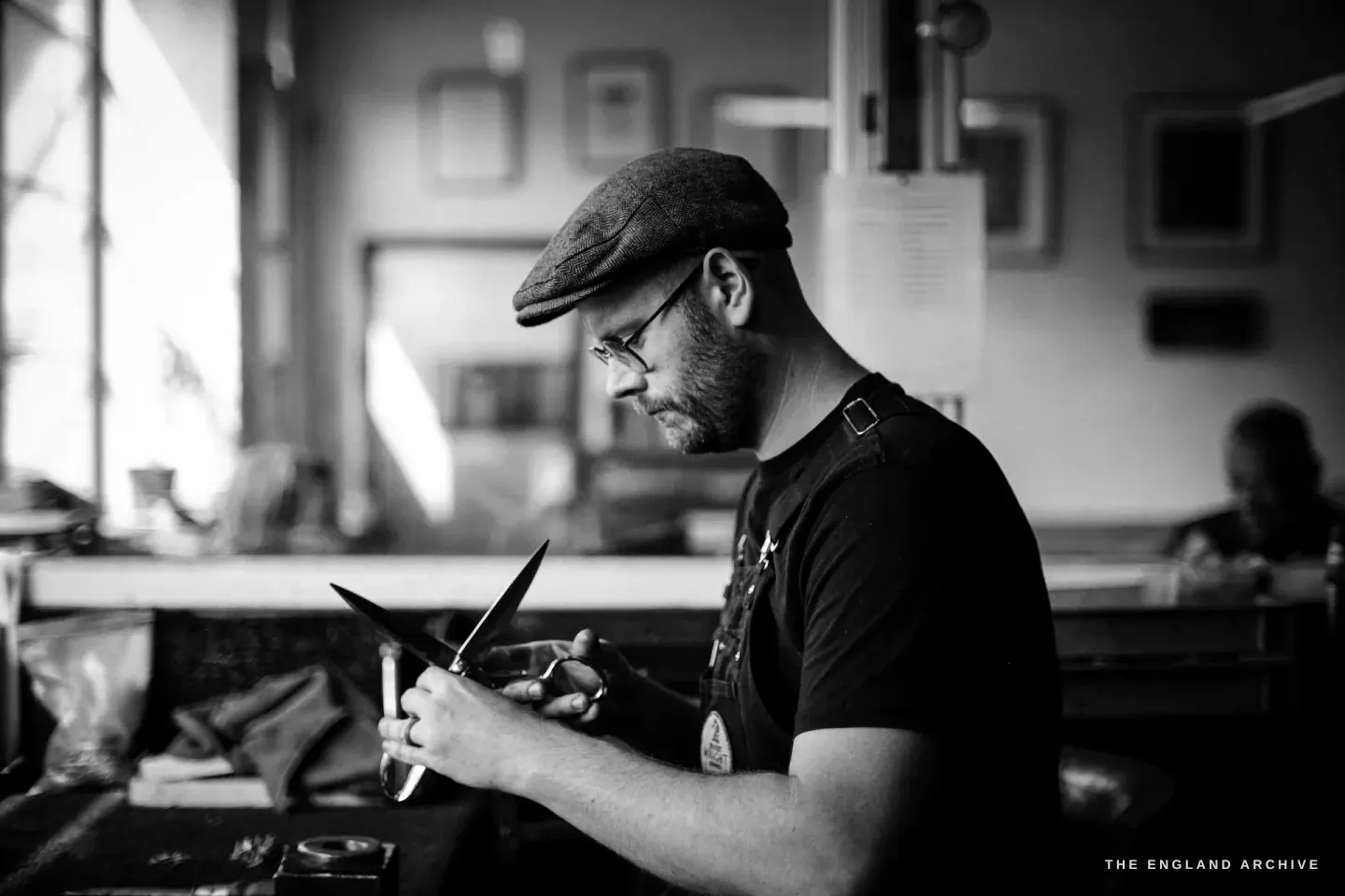 Sam Aston-Clark in a flat cap and leather apron at his bench, working a scissor blade in his hands, soft window light from the left.