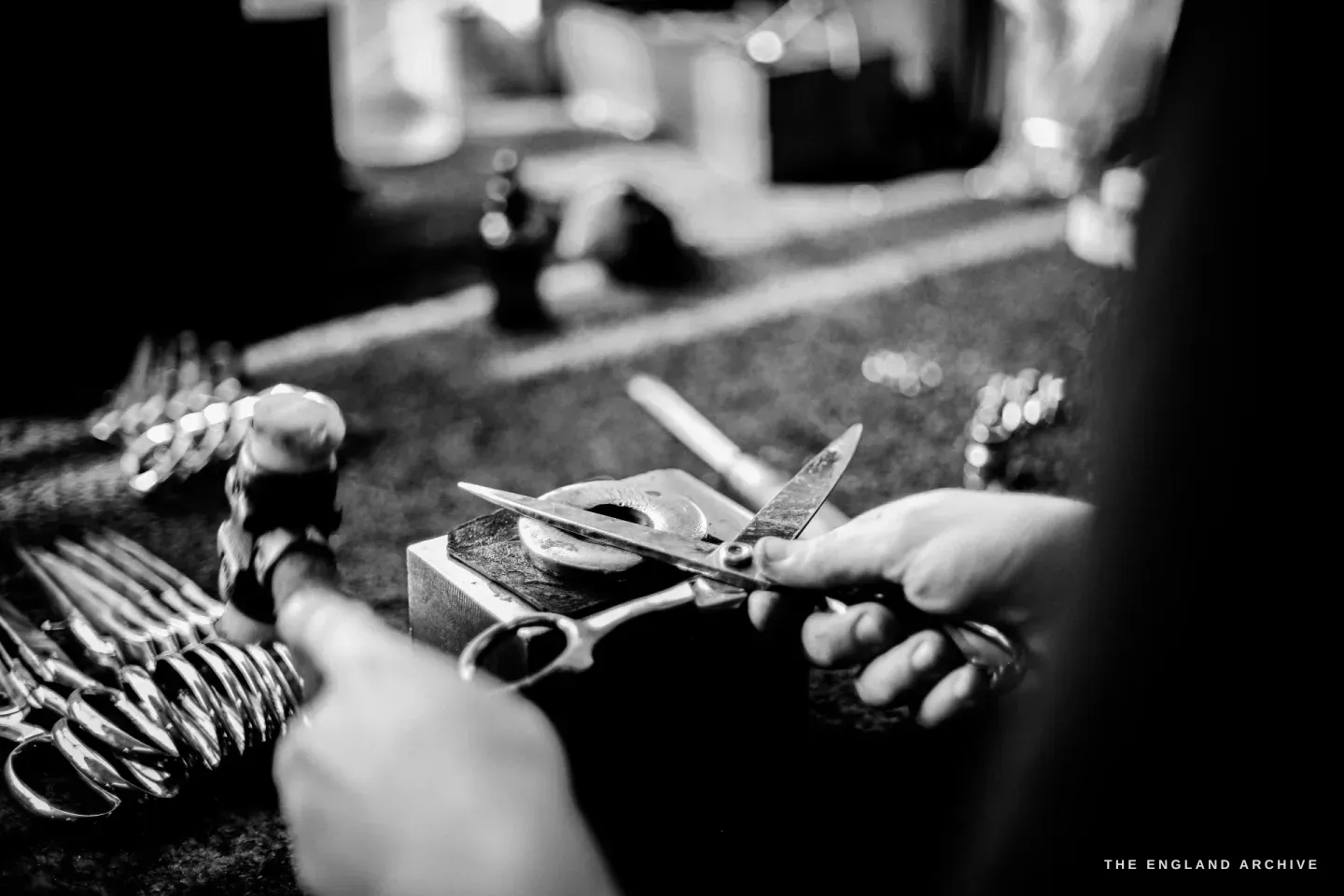 Sam’s hands at a small anvil-stake, a hammer in one hand and a part-made pair of scissors in the other, finished pairs laid out alongside.