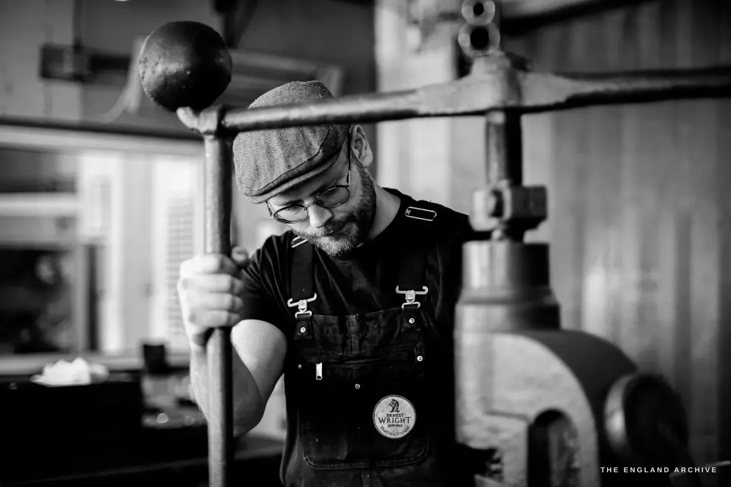 Sam pulling down the long weighted arm of a hand fly-press, head bowed in concentration, the cast frame of the press beside him.