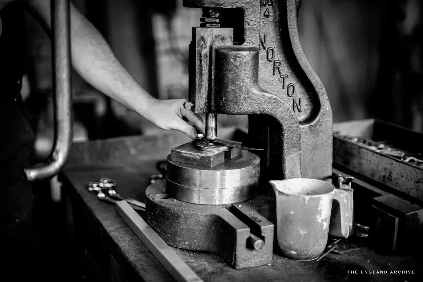 A close view of a cast hand-press marked JOHN NORTON, Sam’s arm guiding a die under the ram, a white jug on the bench.