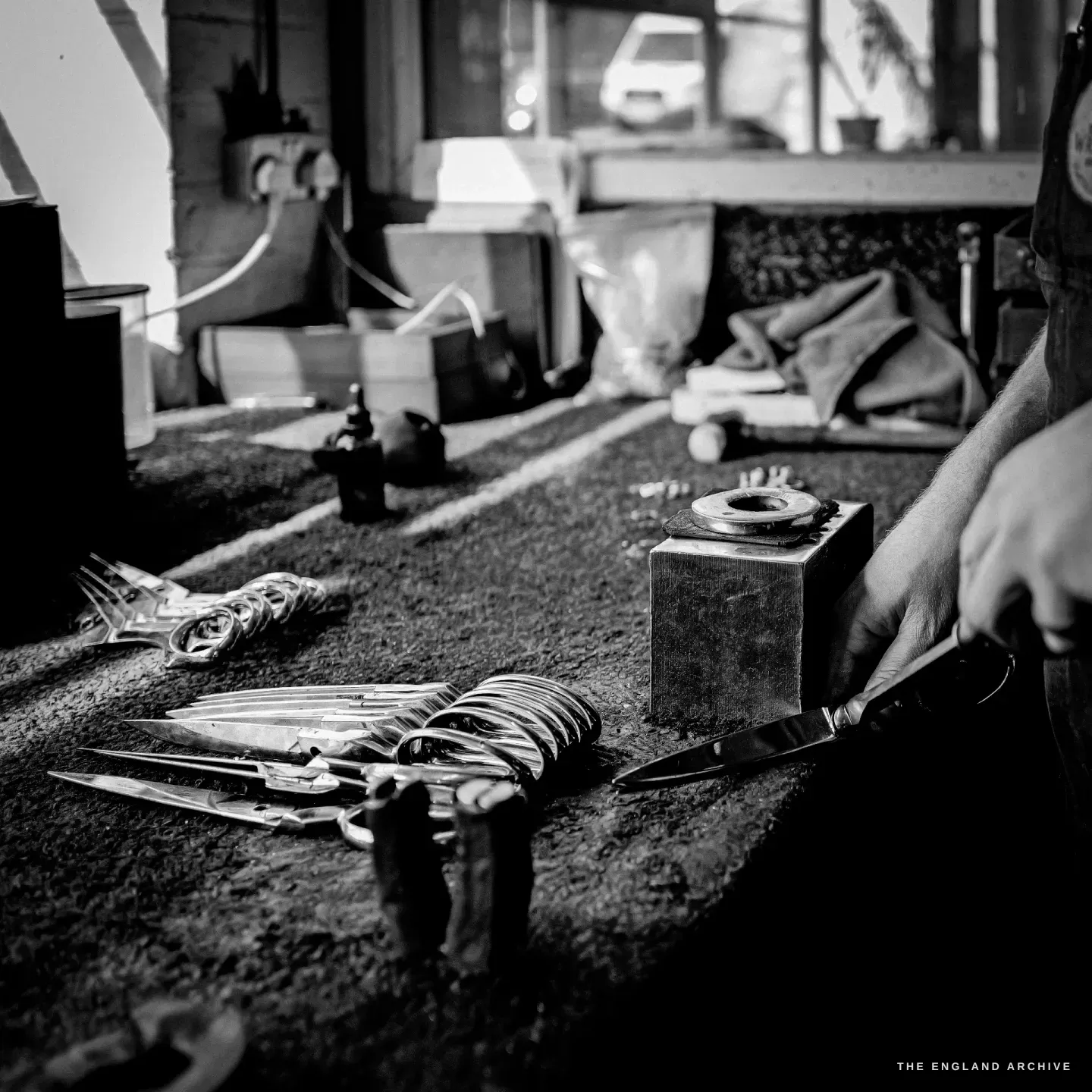 A low view across the bench: rows of part-made scissors laid out, Sam’s hand setting a blade against a small stake block.