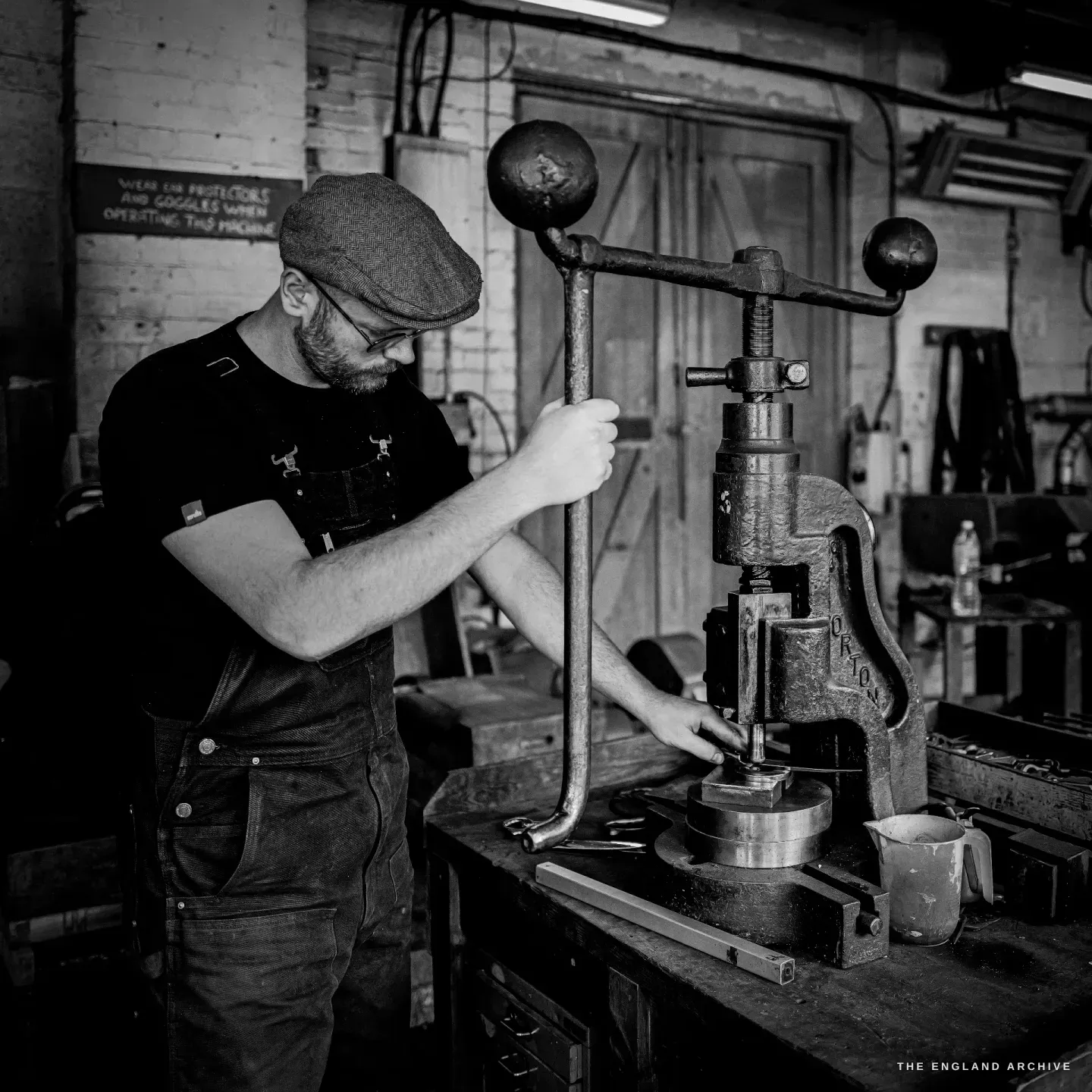 Sam at a large twin-armed fly-press, both hands at the work under the ram, the painted safety sign on the wall behind.