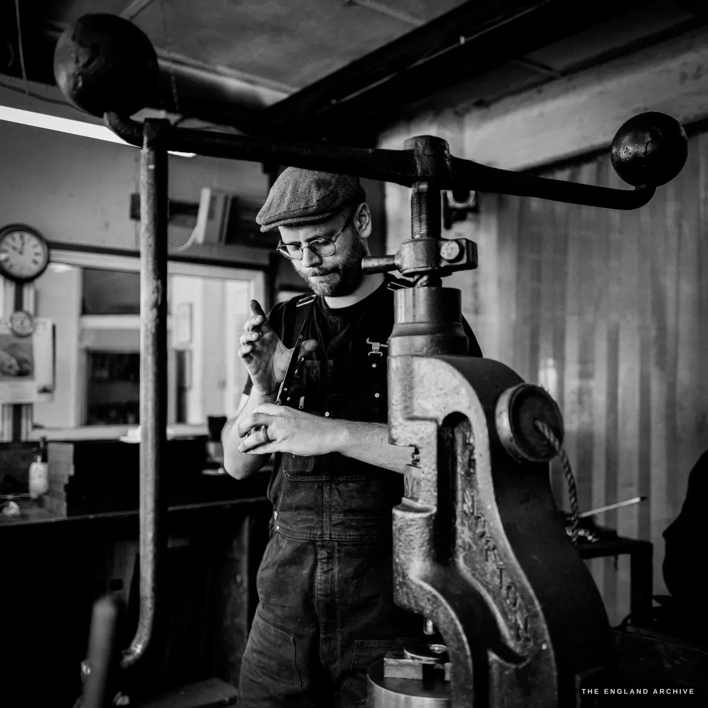 Sam standing at the twin-armed fly-press inspecting a small part in his fingers, a wall clock and framed prints behind.