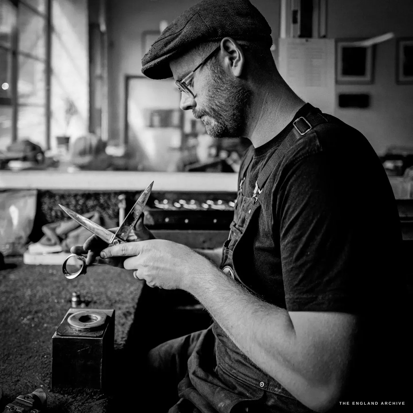 Sam in profile working a blade against a small stake at the bench, finished pairs and a mug beside him.